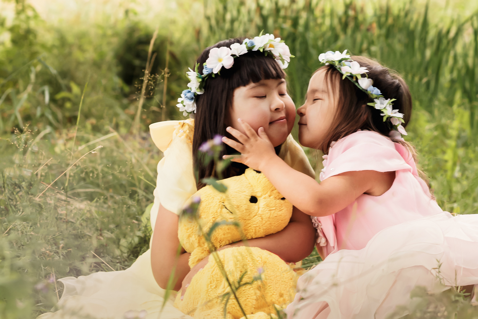 Portrait of two loving sisters wearing princess dresses sitting on a field holding a toy bear.